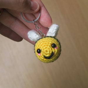 May include: A crocheted bee keychain with a yellow body, white wings, and black eyes. The bee has a smiling mouth and pink cheeks.