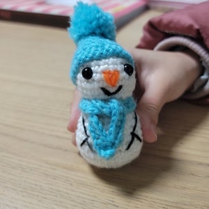 Puede incluir: Un muñeco de nieve tejido a ganchillo con gorro y bufanda azules. El muñeco de nieve tiene ojos negros, una nariz de zanahoria naranja y una boca sonriente. Es blanco con detalles negros y un pompón azul en el gorro.