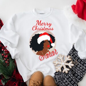 May include: White sweatshirt with a graphic of a Black woman wearing a Santa hat, with the words "Merry Christmas" and "Carol" in red script. The sweatshirt is part of a Christmas theme.