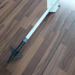 May include: A white and black model rocket with a pointed nose cone and fins. The rocket is made of cardboard and has a black paint job on the nose cone and fins.