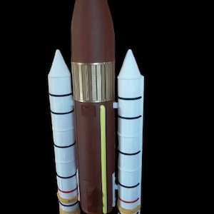 Könnte beinhalten: Ein braunes und weißes Modell eines Raumschiffs mit drei Booster-Raketen. Das Raumschiff ist aus Plastik und hat einen gelben Streifen an der Seite.