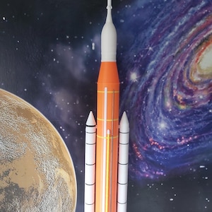May include: A white and orange model rocket with three booster rockets. The rocket is standing on a surface with a space-themed background.