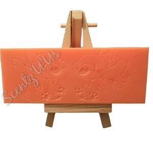 May include: A peach-colored silicone mold with two Stitch characters from the Disney movie Lilo & Stitch. The mold is on a small wooden easel. The text "Scentz' Us" is visible on the mold.