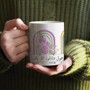 Peut inclure: Mug en céramique blanche avec un motif floral, comprenant des roses violettes et un arc-en-ciel. Le mug porte du texte arabe et est tenu par une personne portant un pull vert.