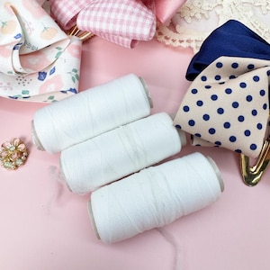 May include: An assortment of craft supplies on a pink surface. The image features white thread spools, decorative hair bows in pink gingham and polka dot patterns, and a floral brooch. The bows have gold-colored clips.