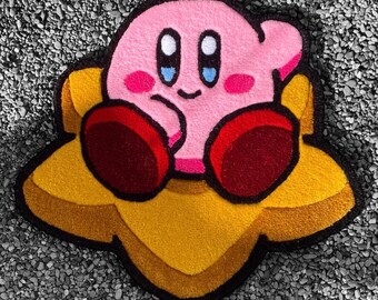 May include: A pink and red plush character with large eyes and a smile, sitting on a yellow star-shaped rug. The character is sitting on the rug with its legs crossed and its arms folded.