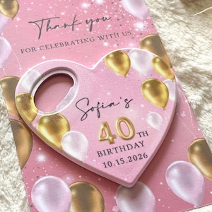 May include: A pink birthday card with gold and white balloons and the text "Thank you for celebrating with us." A heart-shaped bottle opener is on top, with "Sofia's 40th Birthday 10.15.2026" printed on it.