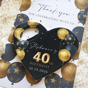 May include: A black heart-shaped magnet with gold accents and the text "Stephanie's 40th Birthday 10.15.2026" is displayed on a card with gold and black balloon graphics. The card reads "Thank you for celebrating with us."
