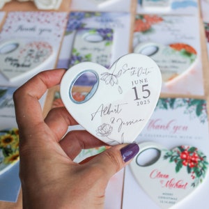 Peut inclure: Un ouvre-bouteille en forme de c&oelig;ur avec une ouverture argent&eacute;e. L'ouvre-bouteille blanc porte l'inscription "Save the Date June 15 2025 Albert & Jessica" avec des motifs floraux. D'autres cartes avec des motifs floraux sont en arri&egrave;re-plan.