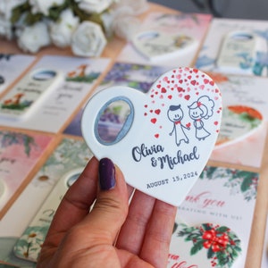 Peut inclure: Un ouvre-bouteille blanc en forme de c&oelig;ur avec un couple de dessins anim&eacute;s, les noms "Olivia & Michael" et la date "15 ao&ucirc;t 2024". Des cartes sur le th&egrave;me du mariage sont visibles.