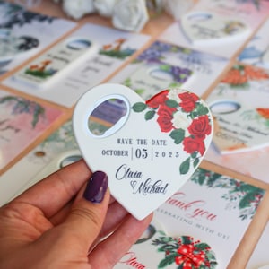 Peut inclure: Un ouvre-bouteille en forme de c&oelig;ur pour mariage, orn&eacute; de roses rouges et blanches et du texte "Save the Date" avec une date et des noms. D'autres cartes avec des motifs floraux sont en arri&egrave;re-plan.