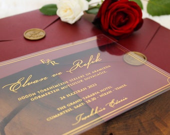 May include: A clear acrylic wedding invitation with gold lettering and a burgundy background. The invitation reads "Elvan and Refik" and includes the date and location of the wedding. The invitation is sitting on a wooden surface.