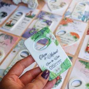 Peut inclure: D&eacute;capsuleur blanc avec un motif floral et de feuilles, avec le texte "Olivia & Michael Save the Date 05 October 2025." D'autres cartes sur le th&egrave;me du mariage sont en arri&egrave;re-plan.