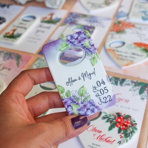 Peut inclure: Un ouvre-bouteille blanc avec un motif floral violet, portant les noms "Alexa & Miguel" et la date "04/05/22". D'autres cartes avec des motifs floraux et en forme de c&oelig;ur sont en arri&egrave;re-plan.