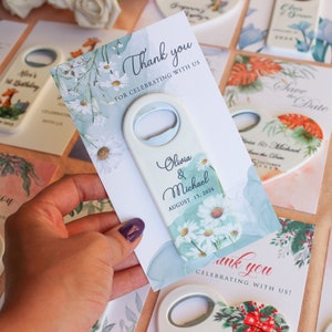 May include: White bottle opener with a floral design and the text "Olivia & Michael August 15, 2024". The bottle opener is on a card with a floral design and the text "Thank you for celebrating with us".