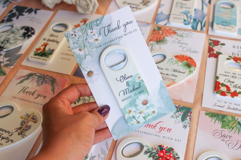 May include: White bottle opener with a floral design and the text "Olivia & Michael August 15, 2024". The bottle opener is on a card with a floral design and the text "Thank you for celebrating with us".
