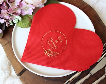 Può includere: Tovagliolo di carta rosso a forma di cuore con scritta dorata "Ena & Dennis 10.09.2021" in un cerchio con un disegno a foglia.