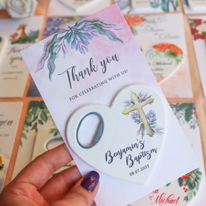May include: A white heart-shaped bottle opener with a cross design and the text "Benjamin's Baptism 08.07.2025". The bottle opener is on a pink card with a floral design and the text "Thank you for celebrating with us!"