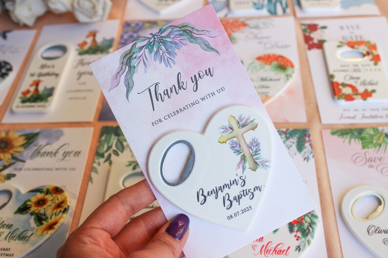 May include: A white heart-shaped bottle opener with a cross design and the text "Benjamin's Baptism 08.07.2025". The bottle opener is on a pink card with a floral design and the text "Thank you for celebrating with us!"