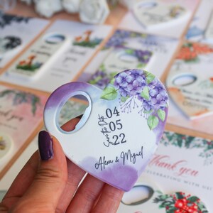 May include: Purple heart-shaped bottle opener with a floral design and the text "Save the Date 04.05.22 Alex & Miguel!"