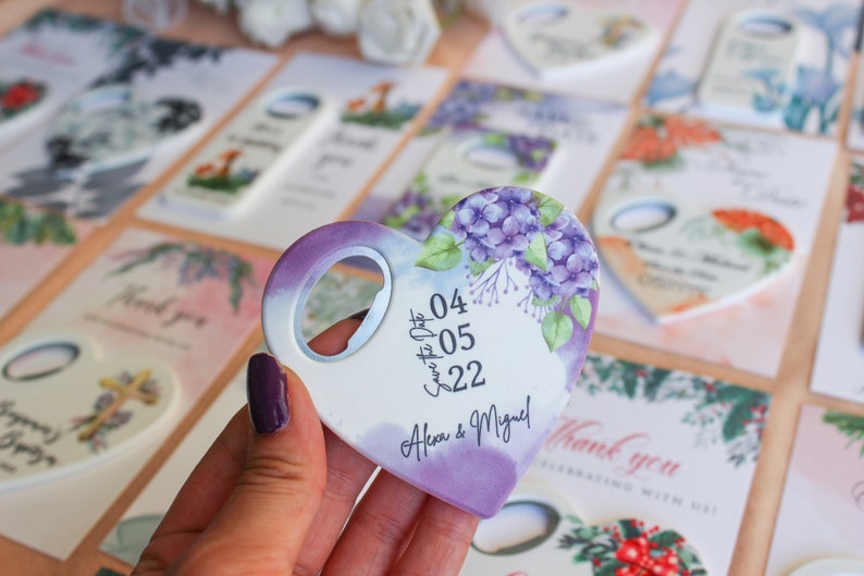 May include: Purple heart-shaped bottle opener with a floral design and the text "Save the Date 04.05.22 Alex & Miguel!"