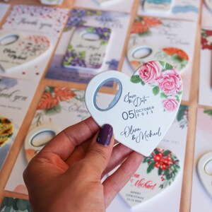 May include: A white heart-shaped bottle opener with a floral design and the text "Save the Date 05|OCTOBER|2025 Olivia & Michael".