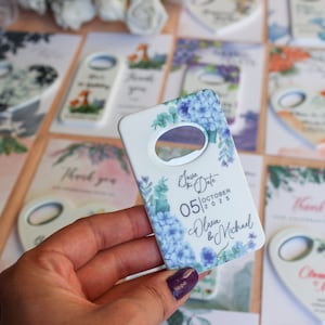 May include: White ceramic bottle opener with a floral design featuring blue and green flowers. The text "Save the Date" is printed on the opener, along with the date "05|OCTOBER|2025" and the names "Olivia & Michael".