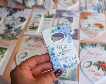May include: White ceramic bottle opener with a floral design featuring blue and green flowers. The text "Save the Date" is printed on the opener, along with the date "05|OCTOBER|2025" and the names "Olivia & Michael".