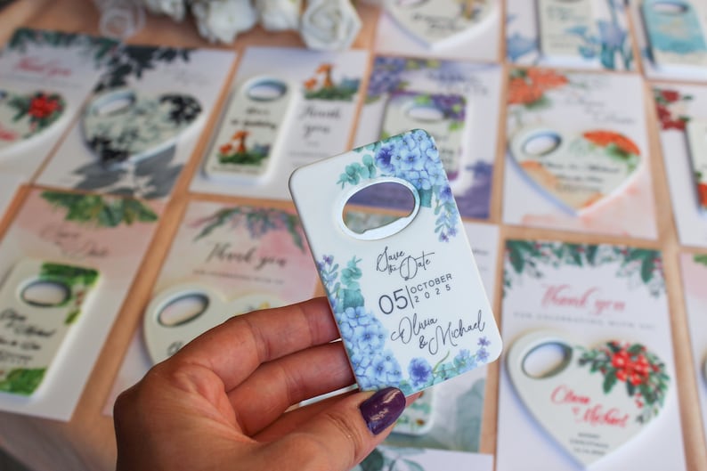 May include: White ceramic bottle opener with a floral design featuring blue and green flowers. The text "Save the Date" is printed on the opener, along with the date "05|OCTOBER|2025" and the names "Olivia & Michael".