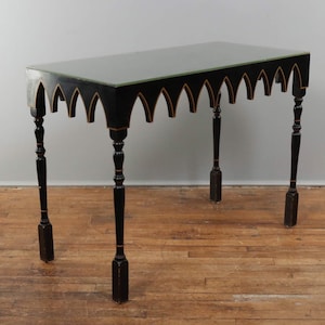 May include: A black console table with a rectangular top and decorative arched trim. The table has four slender, turned legs with gold accents. The tabletop appears to be a dark color, possibly black or dark green.