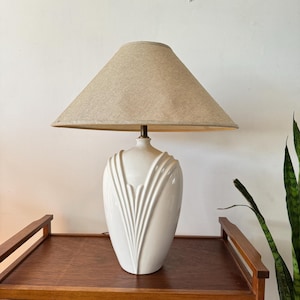 May include: A white ceramic table lamp with a beige lampshade. The lamp base has a decorative, vertical, fan-like design. The lamp is on a wooden table.