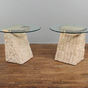 May include: Two side tables with clear glass tops and beige stone bases. The glass tops are semi-circular, and the bases are roughly rectangular with a textured, carved appearance. The tables are set on a wooden floor.