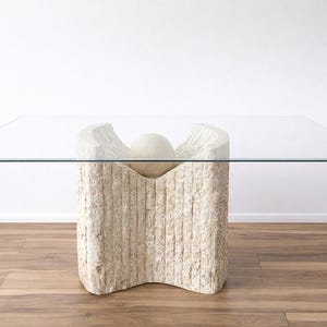 May include: A rectangular glass-top coffee table. The base is made of a textured, off-white stone material, with a rounded, carved design. The table is approximately 48 inches long and 30 inches wide.