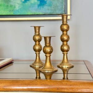 May include: Three gold candle holders with a textured, hammered finish. Each holder has a round base and a round top with a small lip. The holders are arranged in a row on a mirrored surface.