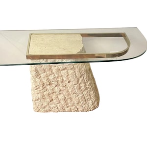 May include: A glass-top coffee table with a unique, textured stone base. The base is a light beige color and has a rough, uneven surface. The glass top is clear and has a brass trim around the edge.