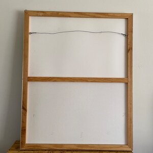 May include: The back of a framed canvas painting with a wooden frame. The frame has a wire attached to the top for hanging.