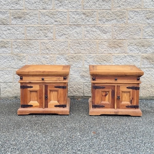 May include: Two wooden nightstands with two doors each. The nightstands have a dark wood finish and metal accents. The nightstands are sitting in front of a brick wall.