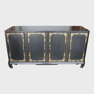 May include: A black sideboard cabinet featuring four doors with gold-colored floral detailing along the edges. The piece has a rectangular top surface, vertical gold-colored handles on each door, and curved feet at the base of the structure.