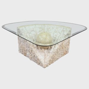 May include: A unique coffee table with a clear glass top. The base is made of textured, light-colored stone, featuring a central sphere. The table has a modern, artistic design, suitable for living rooms.