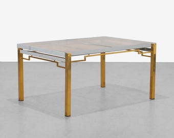 May include: Rectangular table with a glass top and a metal frame. The frame features gold-colored legs and a silver-colored border around the glass. The table's design is geometric and modern, suitable for a living room or dining area.