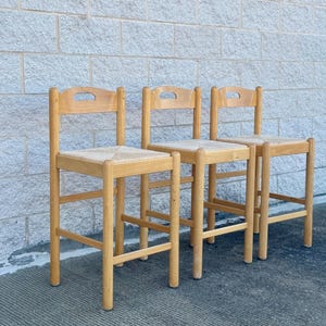 May include: Three wooden chairs with light-colored woven seats and arched backrests. The chairs are made of light-colored wood and have a simple, classic design. They are arranged in a row against a textured, light gray brick wall.