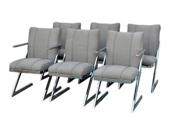 May include: Six gray upholstered armchairs with chrome frames. The chairs have a rectangular back and seat cushions, with a horizontal seam detail. The armrests are chrome and extend outward. The legs are a zig-zag design.