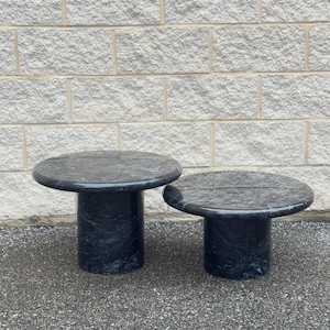 May include: Two black marble side tables with round tops and cylindrical bases. The tables are different sizes and are sitting on a gray concrete surface.