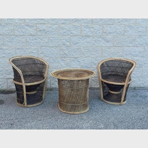 May include: A set of vintage wicker furniture, including two armchairs and a round side table. The chairs have dark brown seats and backs with a light brown frame. The table is cylindrical with a woven top and sides.