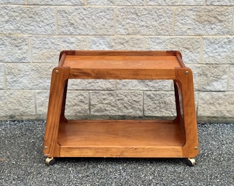 May include: A two-tiered wooden serving cart with a light brown finish. The cart has a rectangular top and bottom shelf, and it is supported by four legs. The cart has four brass wheels.