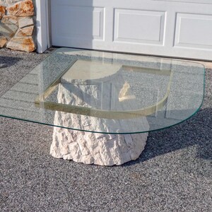 May include: A glass-top coffee table with a unique, rounded, stone base. The base is made of white and beige stone and has a rough, textured surface. The glass top is clear and has a brass trim.