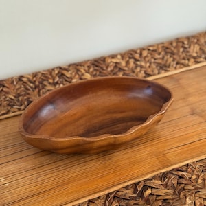 May include: A wooden oval-shaped bowl with a scalloped edge. The bowl is made of a warm-toned wood and sits on a woven surface. The bowl's interior is smooth and polished, showcasing the natural wood grain.