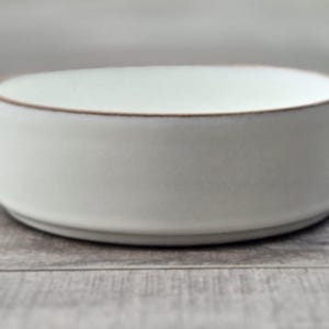 以下が含まれることがあります： 茶色の縁が付いた、小さくて丸いオフホワイトのセラミックボウル。ボウルはシンプルでモダンなデザインで、灰色の木製の表面に置かれています。ボウルの寸法は、高さ約5cm、直径約13cmです。