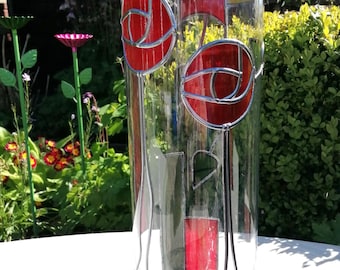 May include: A clear glass vase with red and green stained glass floral designs. The vase features two large red rose-like shapes and smaller rectangular and leaf-shaped accents. The vase is set against a blurred background of greenery.