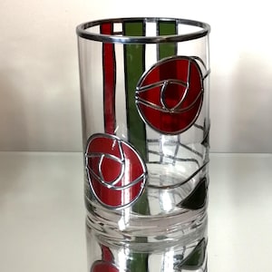 May include: A clear glass vase with a silver rim and decorative red rose designs. The vase has vertical red and green stripes, and the rose designs are outlined in silver. The vase is reflected on a white surface.